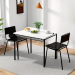 Современная обеденная зона, белый стол, два черных стула, оранжевый подвесной светильник, растение в горшке, серый ковер.