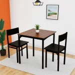 Квадратный обеденный стол, два черных стула, оранжевая стена, растение в горшке, картина на стене.