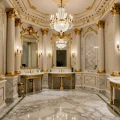 Интерьер ванной в стиле ампир: белые мраморные стены, золотые колонны, большие зеркала в рамах и хрустальная люстра. Роскошный дизайн с классическими элементами и элегантной сантехникой.